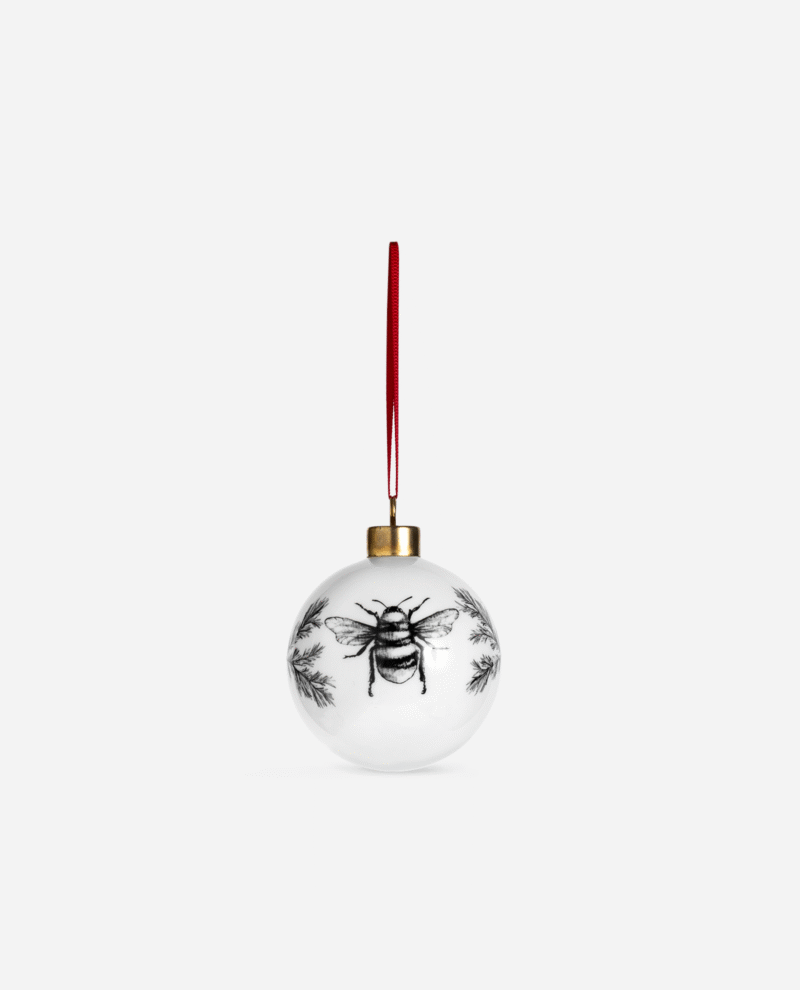 White bone china Manchester Bee Christmas bauble with black illustrated bee design and pine sprigs, hanging from red ribbon