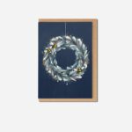 Christmas wreath greeting card with sage green foliage and yellow-striped bees on navy blue background beside kraft envelope