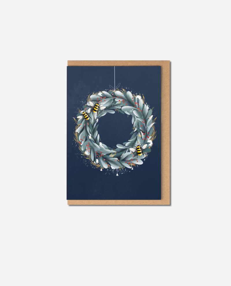 Christmas wreath greeting card with sage green foliage and yellow-striped bees on navy blue background beside kraft envelope