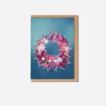 Christmas Wreath Two greeting card by The Sculpts showing circular wreath with pink ribbon, purple foliage, coral pinecones and bee on blue card with brown envelope
