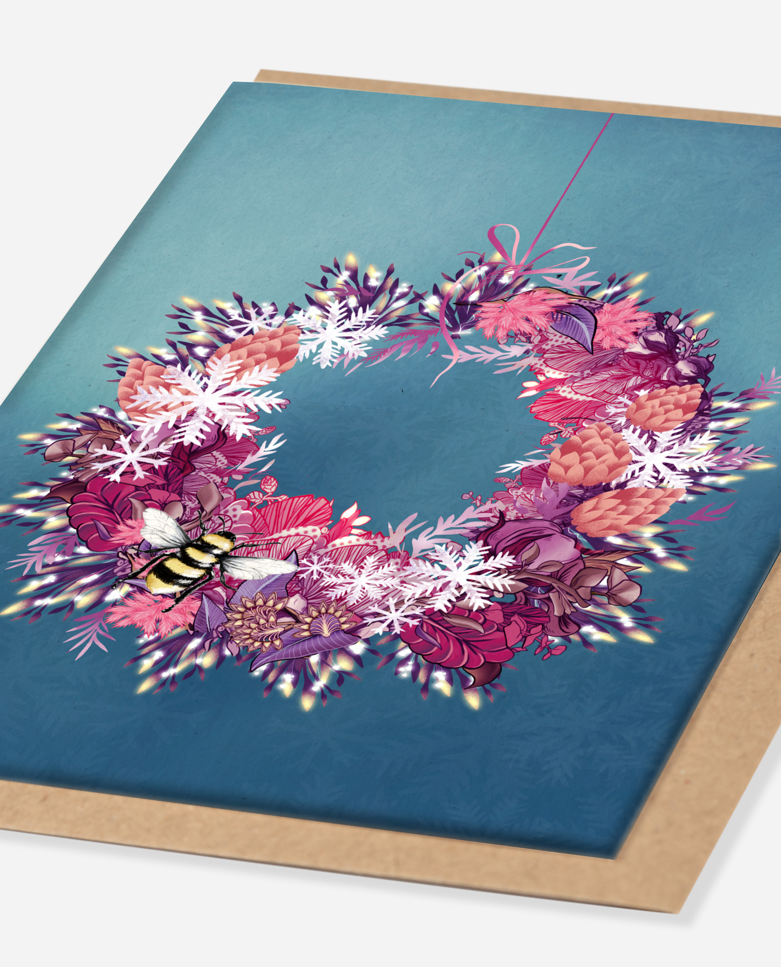 Angled view of Christmas wreath greeting card showing detailed floral illustration with snowflakes, pinecones and bee on teal blue card with kraft envelope