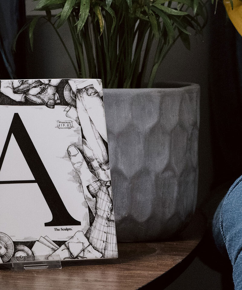Affleck's Palace alphabet tile leaning against grey cushion on yellow chair with potted plant in background