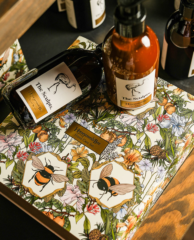 Honeycomb Bee Hand Care Set by The Sculpts, styled with illustrated floral packaging and amber glass bottles in a warm, luxury setting.
