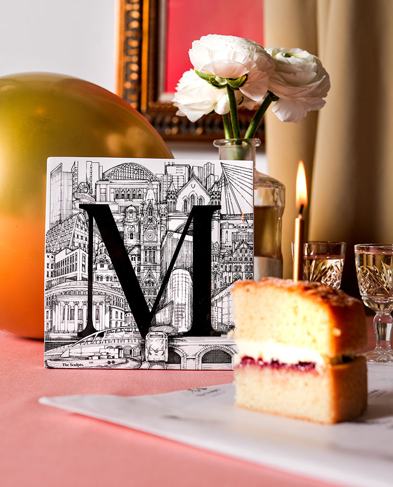 Manchester Letter M tile styled in a luxury festive setting with flowers, candlelight, a gold balloon and a slice of cake on a pink tablecloth, creating a warm celebratory atmosphere.
