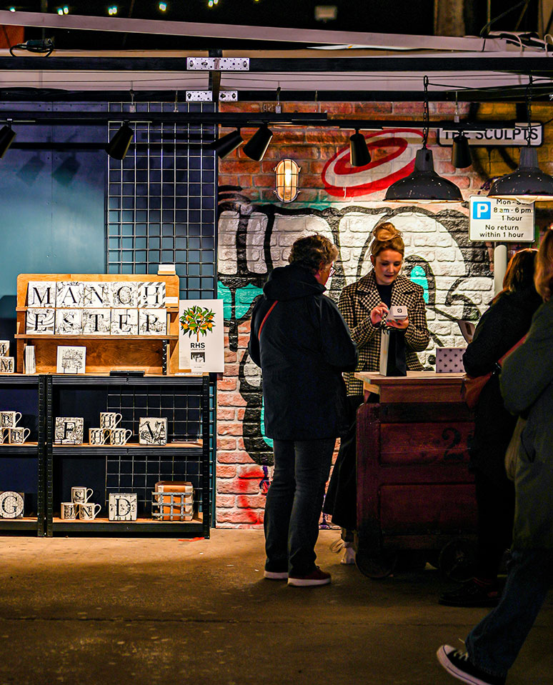 The Sculpts market stall at evening showing gift voucher display with Manchester ceramic homeware and graffiti wall backdrop